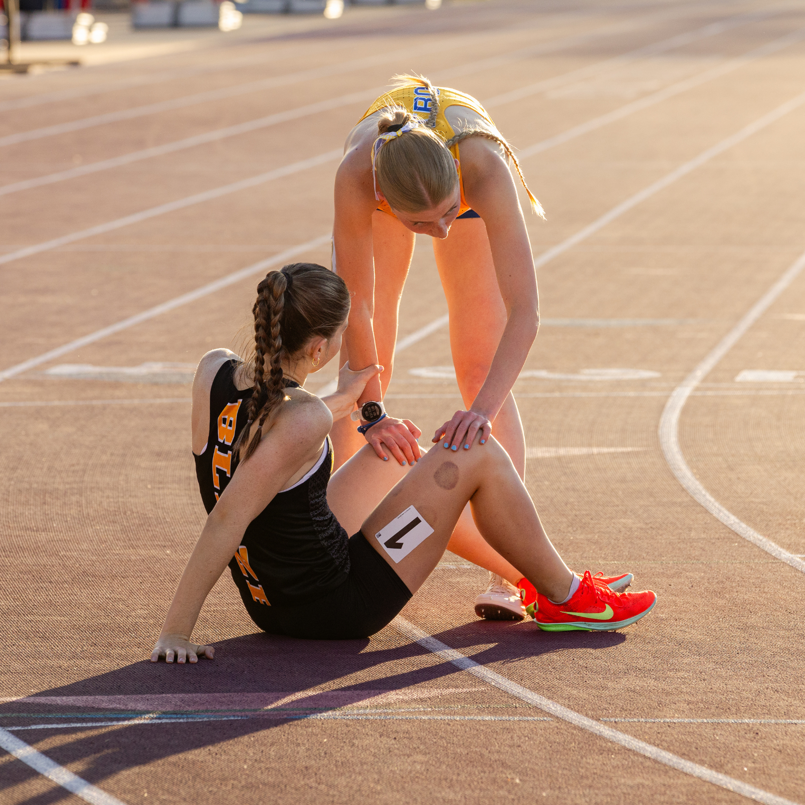 Two athletes on a running track, one helping the other who is sitting.